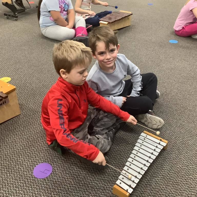 a picture of Two young boys sitting on a classroom carpet; one in a red sweatshirt holds a mallet over a small glockenspiel while the other boy smiles at the camera