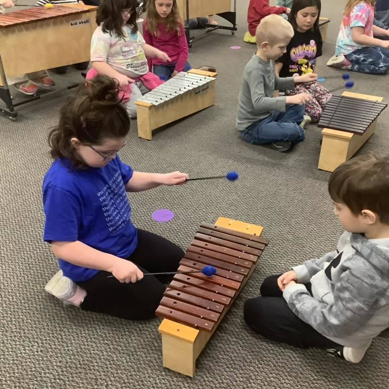 a picture of Based on the image you have selected in your Thrillshare uploader, here are a few options for the alt text:  Option 1: Detailed & Descriptive (Best for accessibility)  A young girl in a blue shirt and glasses sits on the floor, focused on playing a wooden xylophone with blue mallets. Other students are visible in the background, also sitting on the carpet and playing instruments