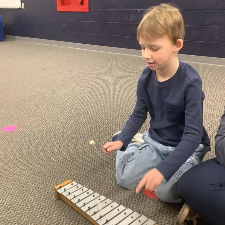 picture of Two young boys sitting on the floor of a music room, focused on playing a wooden xylophone. One boy is wearing a black Under Armour sweatshirt and the other is wearing a grey t-shirt and glasses
