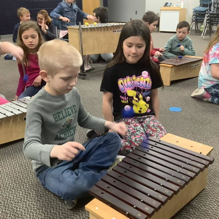 a picture of A boy in a blue "Believe" sweatshirt and a girl in a black "Believe" t-shirt sit on the floor of a music room, focused on playing a large wooden xylophone with mallets