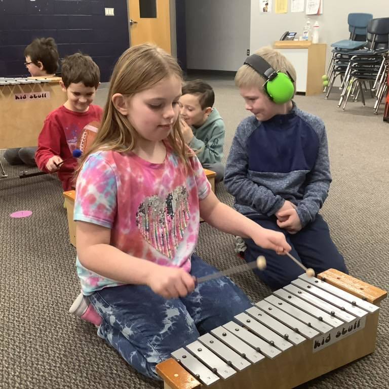 picture of A young girl in a tie-dye shirt plays a wooden xylophone while two boys watch closely; one boy in a blue sweatshirt is wearing green noise-canceling headphones