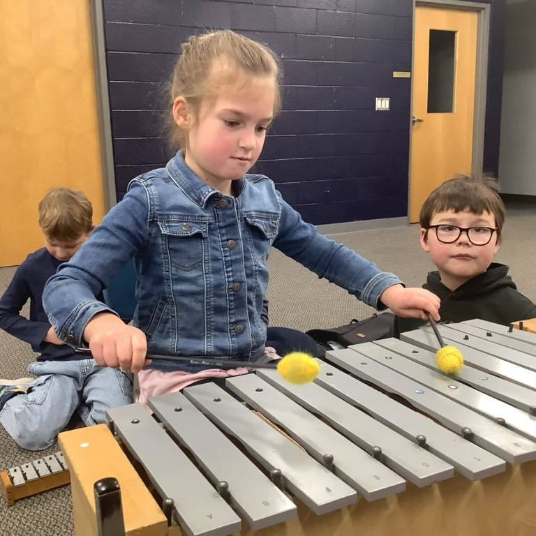 picture of A young girl in a denim jacket and a boy with glasses sitting on the floor, using yellow mallets to play a large silver metallophone