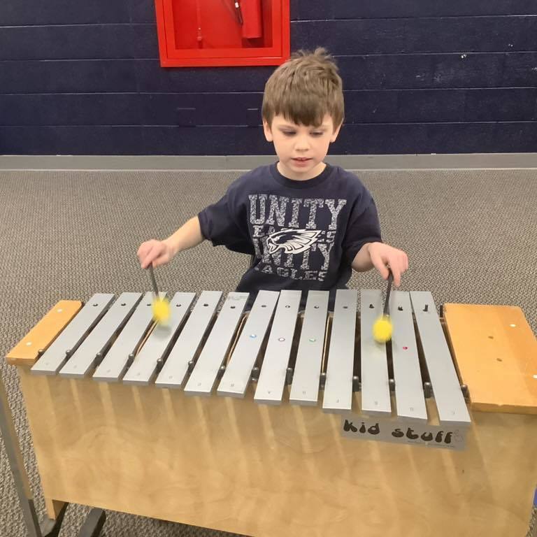picture of A young boy wearing a navy "Unity Eagles" t-shirt stands at a large silver metallophone, holding a mallet in each hand and focusing on the metal bar