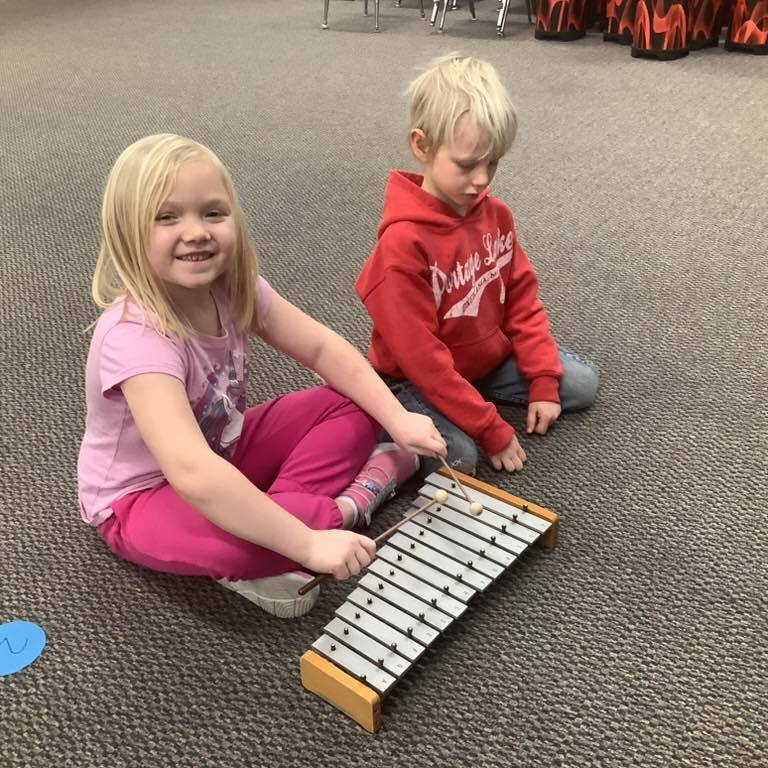 picture of A young girl in a pink shirt and a boy in a red "District 506" hoodie sitting on the floor together, practicing on a wooden xylophone