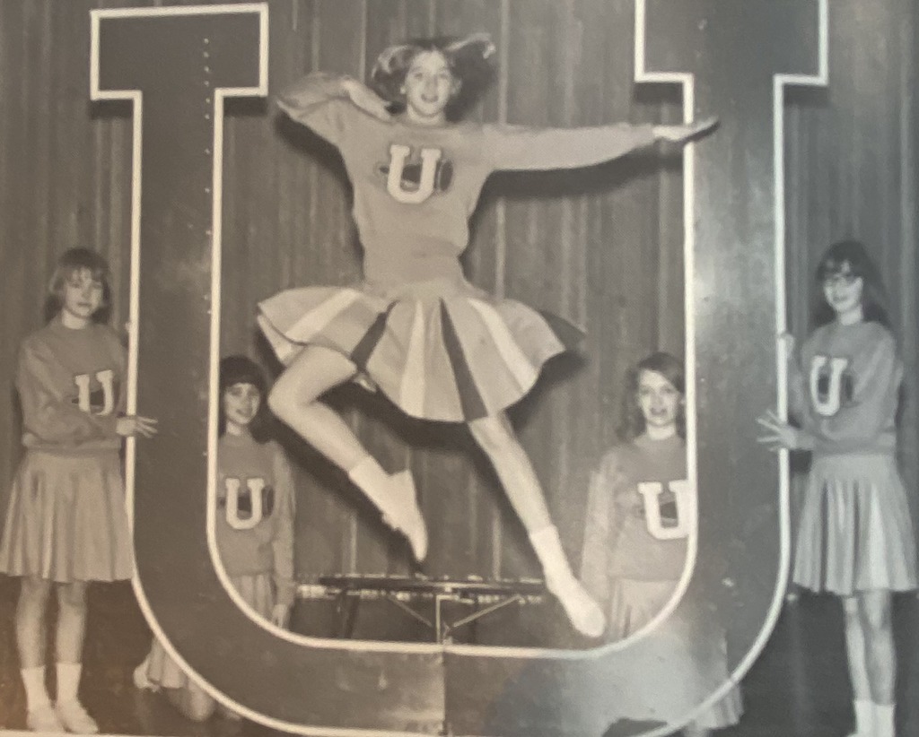 picture of A vintage black-and-white photo of four cheerleaders posing with a large cutout of the letter "U." The cheerleader in the center is captured mid-jump in a dynamic split, while the others stand behind the "U" holding pompoms