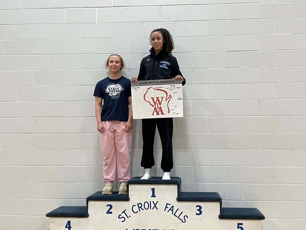 Two female wrestlers on a victory podium at St. Croix Falls. The athlete on the first-place spot holds a Regional Championship bracket, and the athlete on the second-place spot stands beside her.