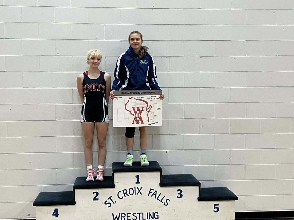Two female wrestlers posing on the victory podium, with the regional champion on the top tier holding the WIAA bracket board.