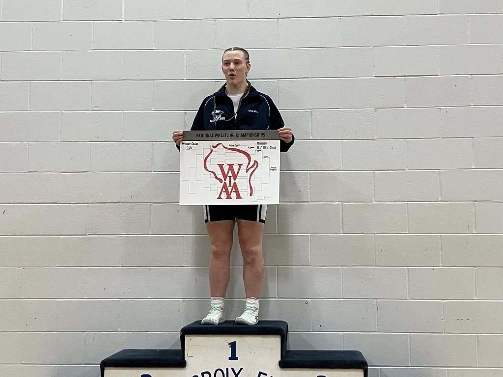 A female wrestler standing alone on the first-place spot of a victory podium, holding a Regional Wrestling Championship bracket board.