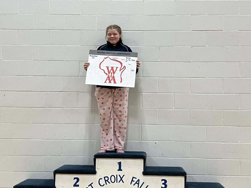 A female wrestler standing alone on the first-place spot of the St. Croix Falls podium, holding her Regional Championship bracket board