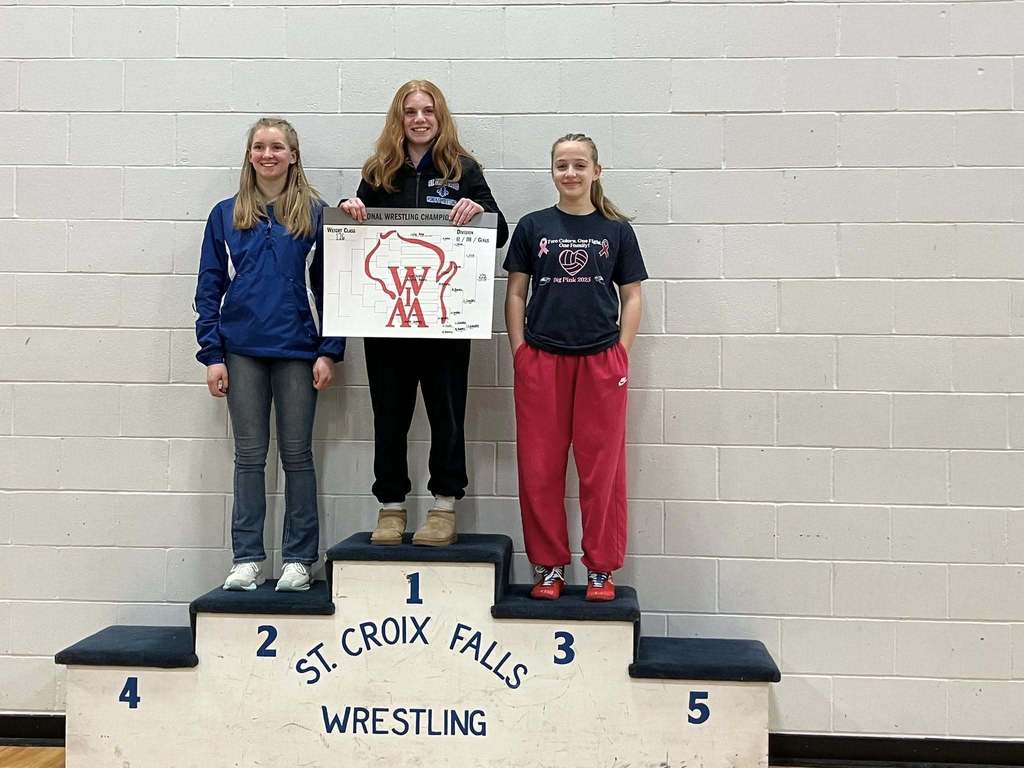 Three female wrestlers posing on a victory podium. The athlete in the center stands on the first-place spot holding a championship bracket, flanked by the second and third-place finishers
