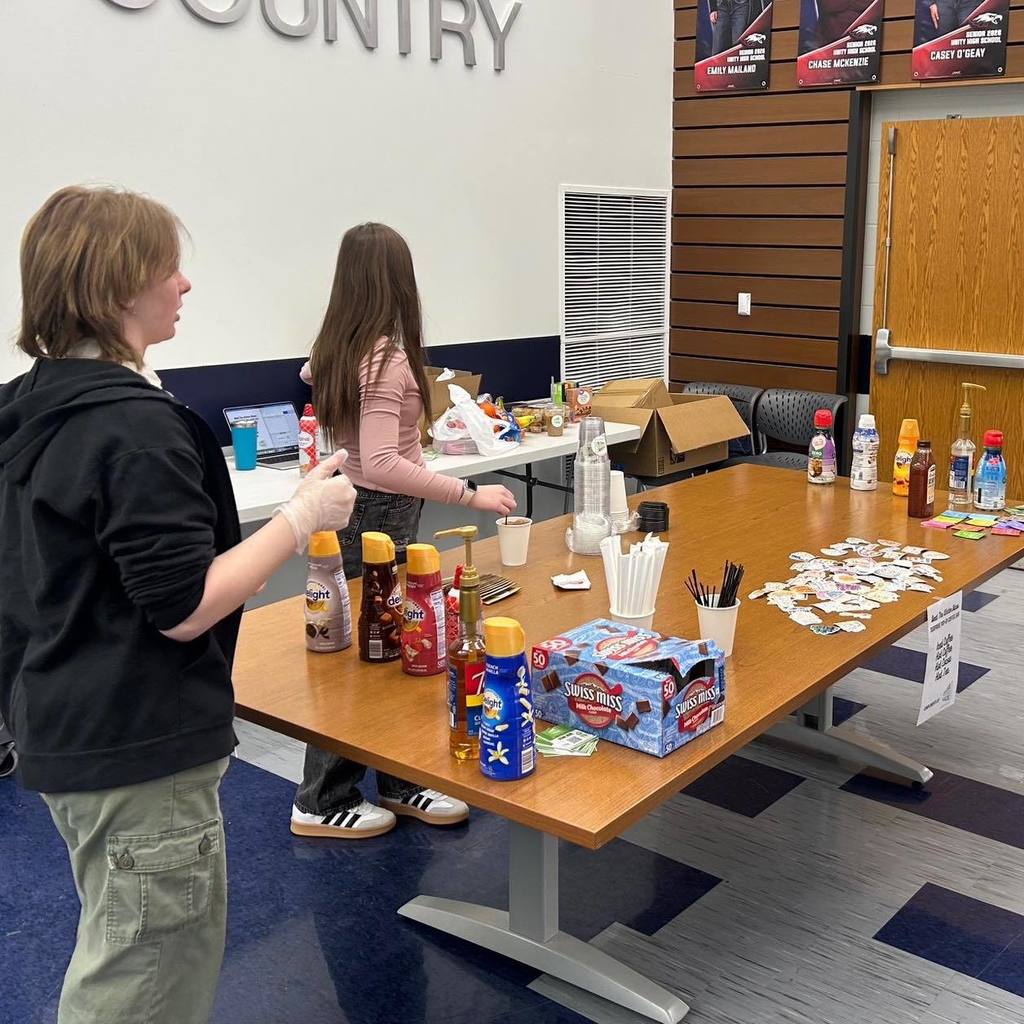 For the fourth and final image in your post, which shows the full table setup from a side angle, here are your options:  Option 1: Balanced A side view of the Unity coffee bar table featuring various syrups, creamers, and boxes of Swiss Miss hot cocoa, with students and staff interacting in the background