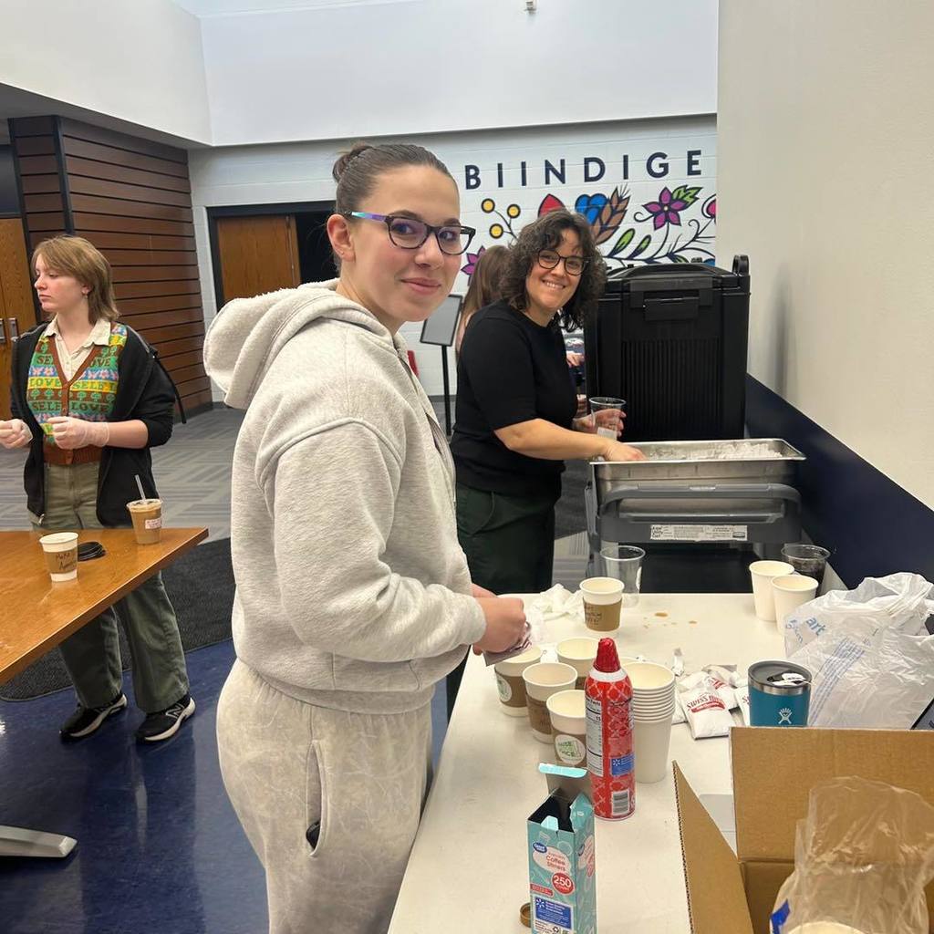 For the third image in your sequence—the one showing the student at the table with the "BIINDIGE" sign in the background—here are your alt text options:  Option 1: Balanced A Unity student in a grey hoodie preparing a drink at the coffee bar, with a colorful "BIINDIGE" sign and flower mural in the background