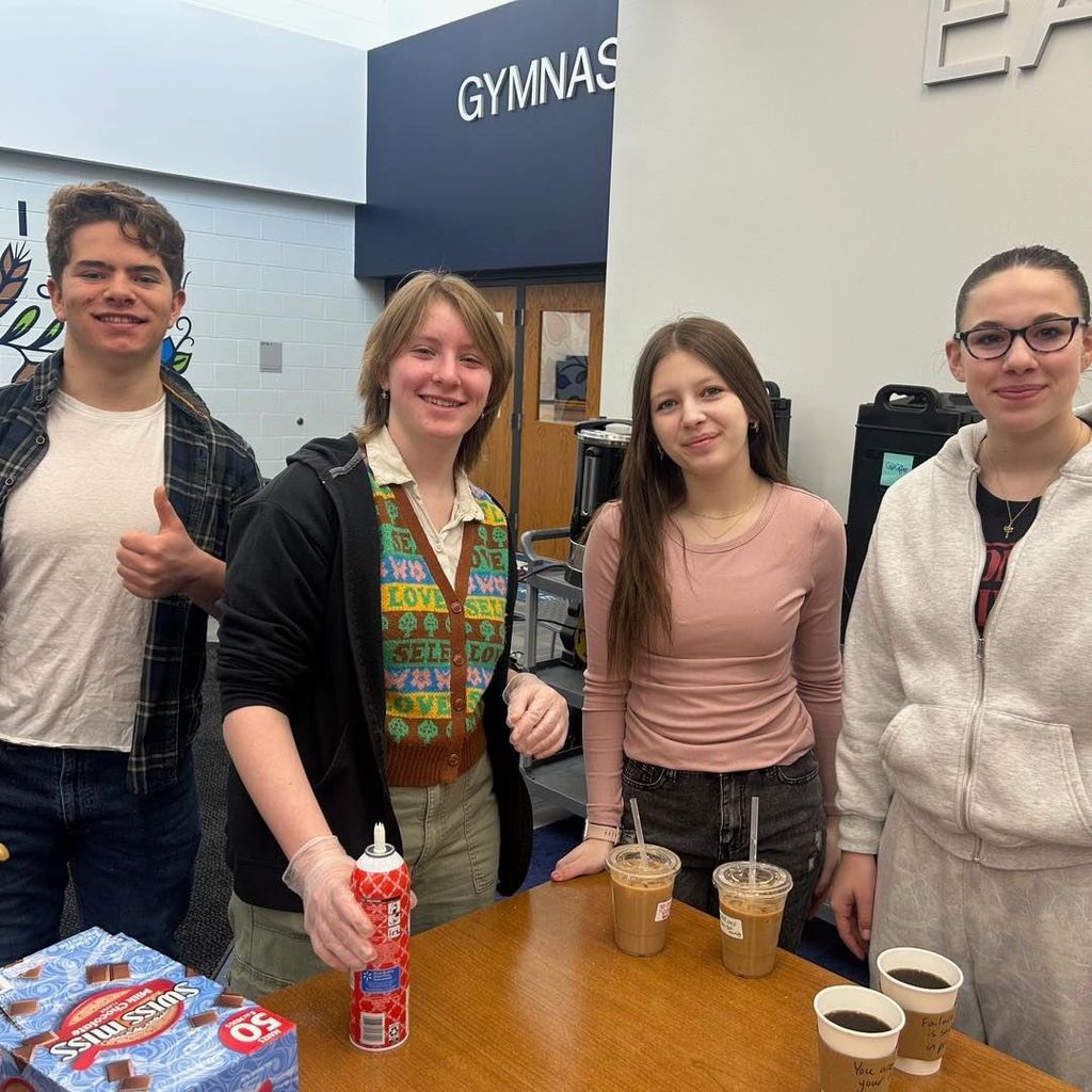 Based on the image you're uploading and the context of your post, here are a few options for your alt text, ranging from concise to more descriptive:  Option 1: Balanced (Recommended) Four smiling Unity students standing behind a table with coffee, tea, and hot cocoa supplies in the high school hallway near the gymnasium