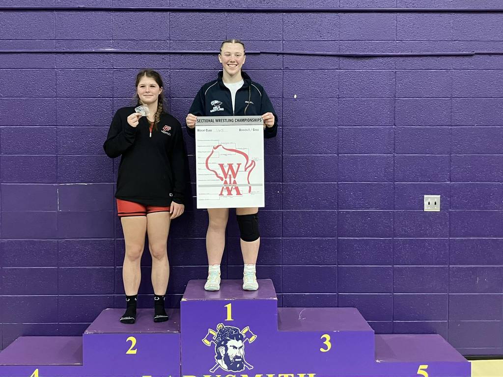 For this image, a short and effective alt text would be:  "Two female wrestlers standing on a podium. The wrestler in the first-place spot is smiling and holding a 'Sectional Wrestling Championships' bracket board featuring the WIAA logo