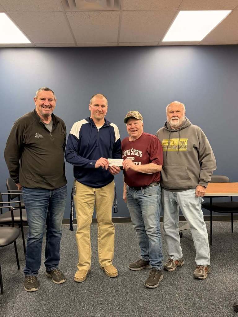 Four men standing in an office setting, smiling and holding a donation check. One man in a maroon shirt representing Youth Sports Unlimited is handing the check to a man in a blue and white pullover.