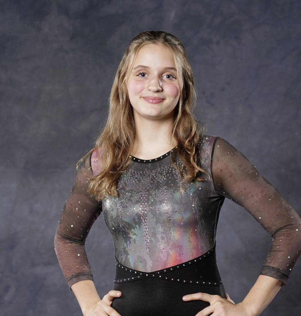 Portrait of Unity student Cami Korf smiling in a sparkly grey and black gymnastics leotard against a grey studio background.
