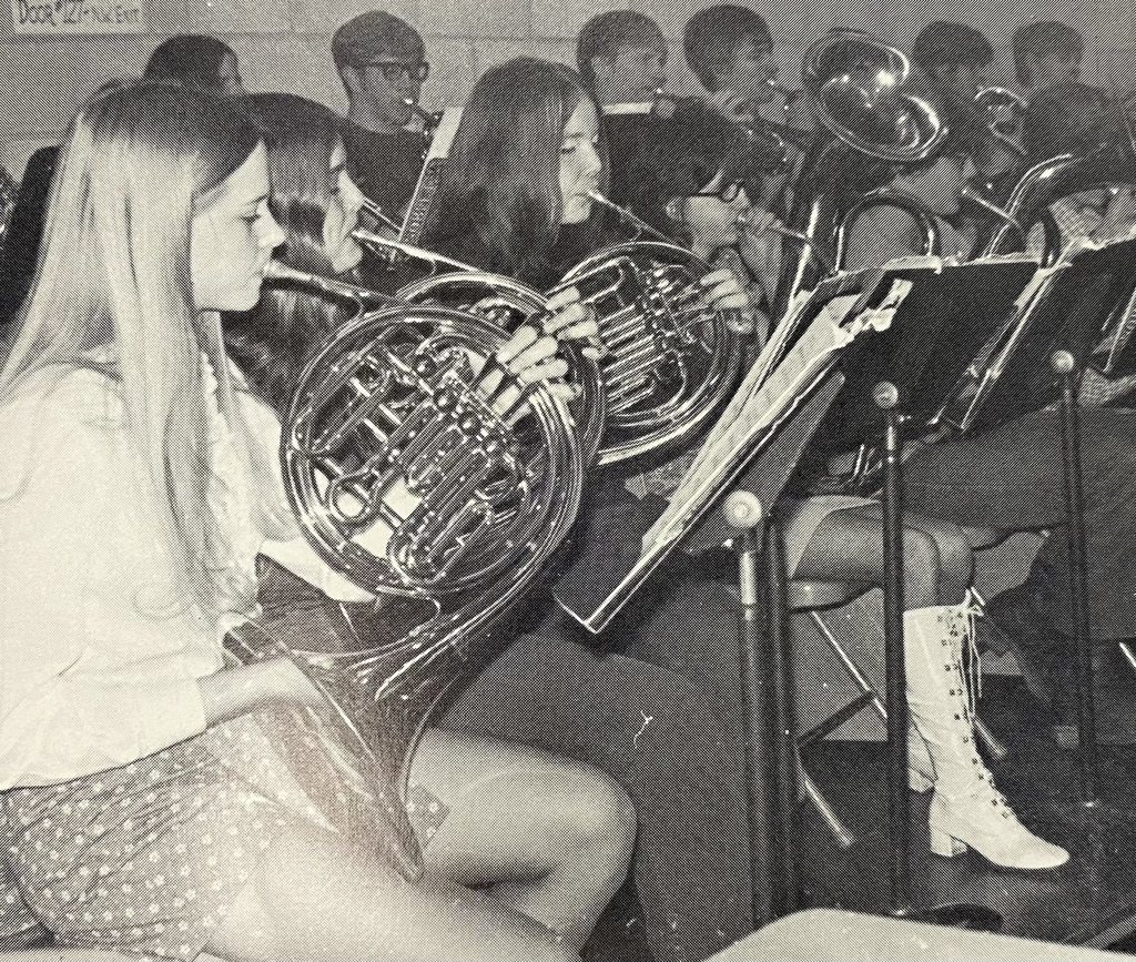A vintage black and white photograph of several students in a school band playing French horns during a performance. The students are dressed in 1970s-style clothing, including one student in the foreground wearing tall white lace-up boots.