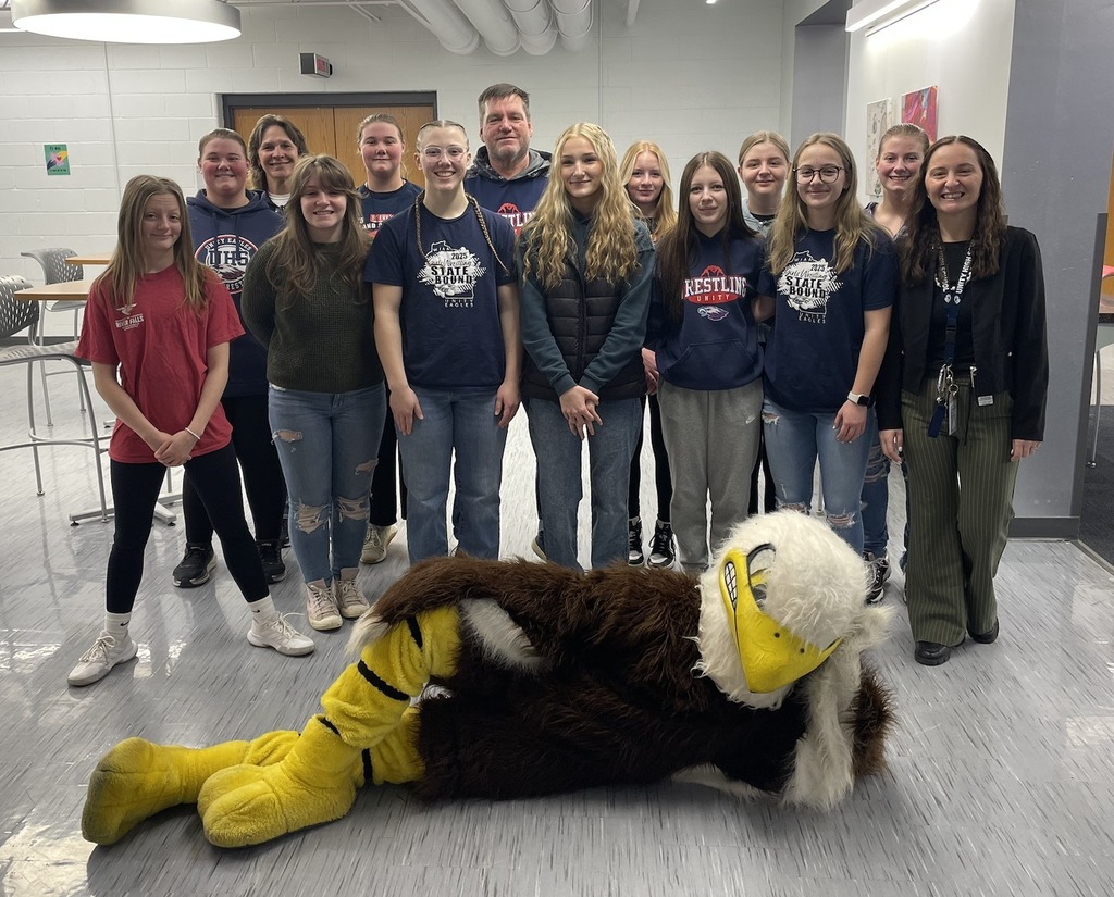 A large group of Unity students and a staff member posing together in a school hallway. In the foreground, the school's eagle mascot is playfully laying on the floor in front of the group.