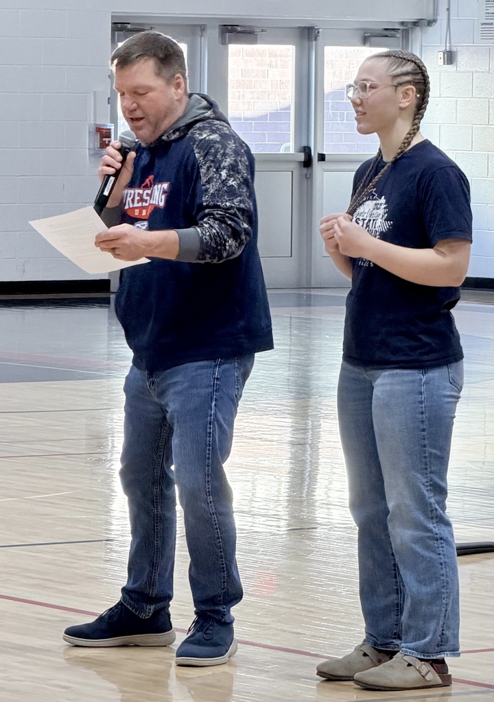 Coach Anderson, wearing a navy and grey Unity Wrestling hoodie, speaks into a microphone while standing next to student Evelyn Fugate in the school gymnasium.