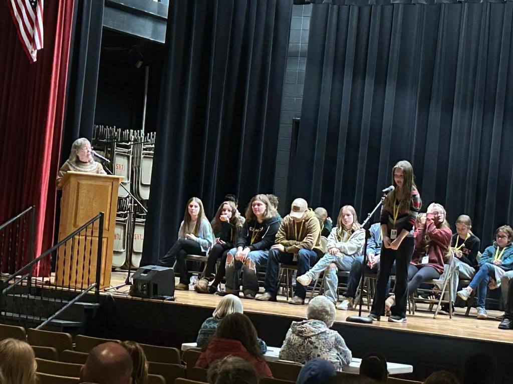 A young female student stands at a microphone on a stage during the CESA Regional Spelling Bee, with other contestants seated behind her.