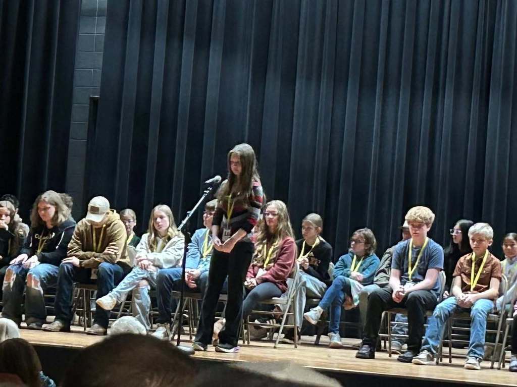 A young female student stands at a microphone on a stage during the CESA Regional Spelling Bee, with other contestants seated behind her.