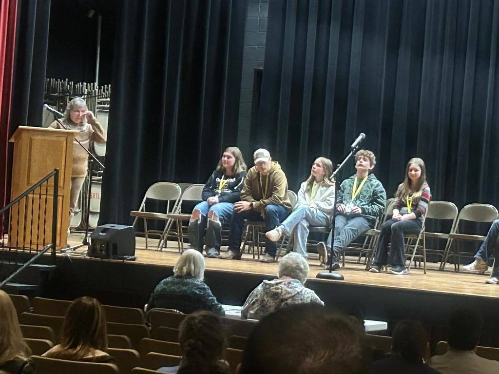 A wide shot of the CESA Regional Spelling Bee stage, showing several student contestants seated in a row and a moderator at the wooden podium to the left.