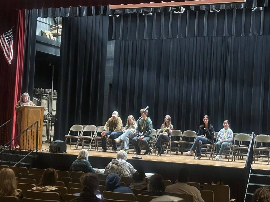 A wide shot of the CESA Regional Spelling Bee stage, showing several student contestants seated in a row and a moderator at the wooden podium to the left.