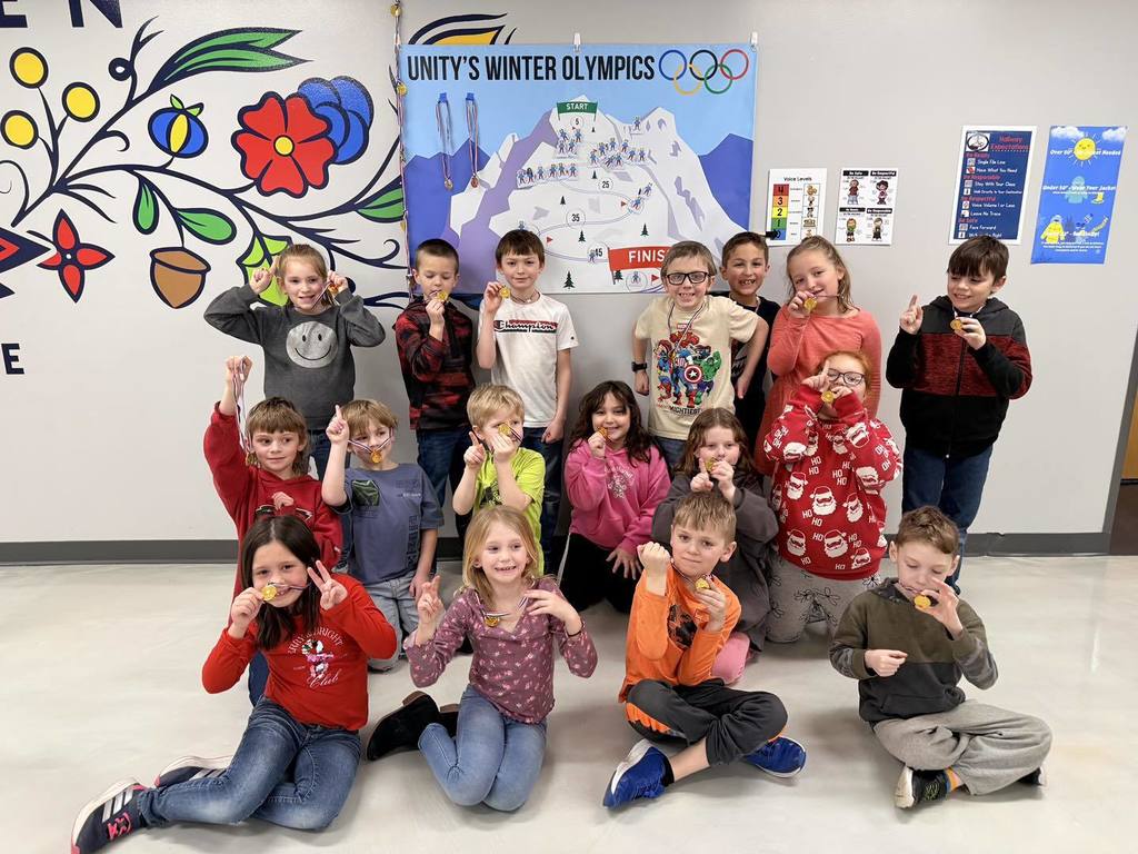 group of elementary students sitting on the floor in a classroom, smiling and holding up small medals, in front of a colorful "Unity's Winter Olympics" poster.
