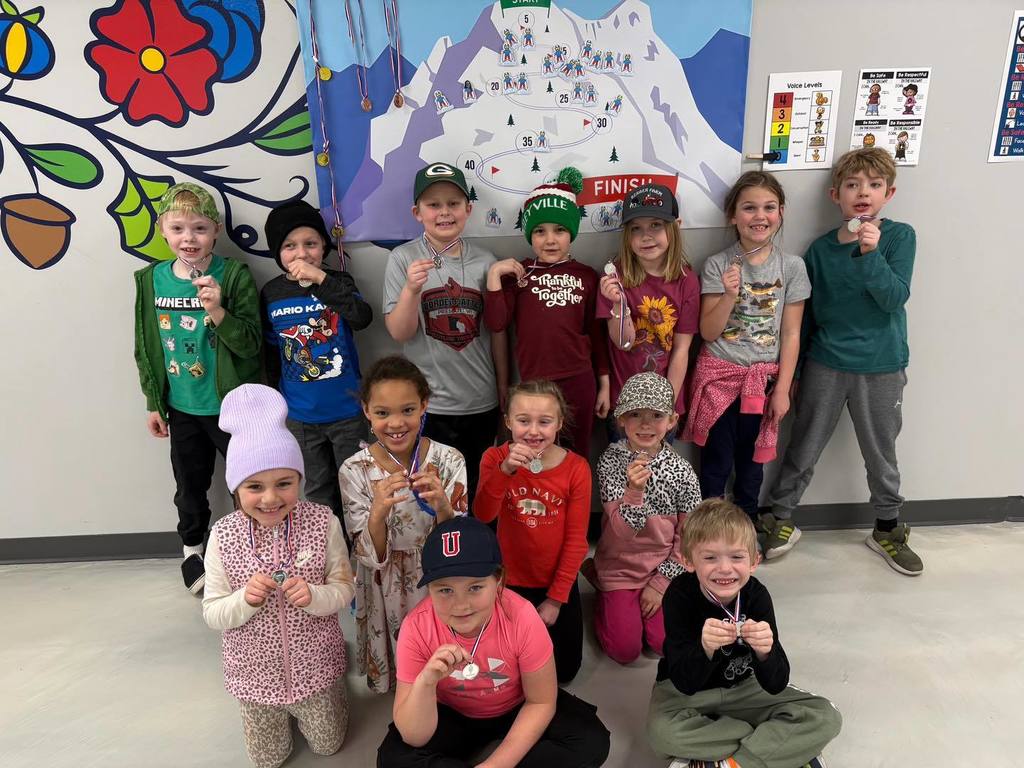 A group of first-grade students from Mrs. Grams’ class posing together indoors. Several students are smiling and proudly holding up silver medals. In the background, there is a large colorful wall display featuring a mountain, flowers, and a 'Finish' line for their Winter Olympics activity.