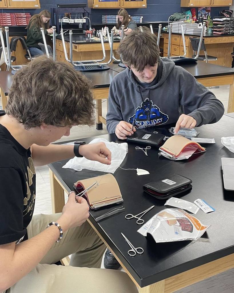 Two high school students sitting at a lab table, focused on practicing suturing techniques on medical training pads. They are using surgical tools and thread as part of the 'Suturing 101' station.