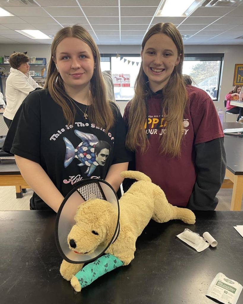 Two high school students smiling while posing with a yellow lab plush toy wearing a veterinary recovery collar (cone). They are participating in a hands-on Small Animals vet science lab at Unity High School.
