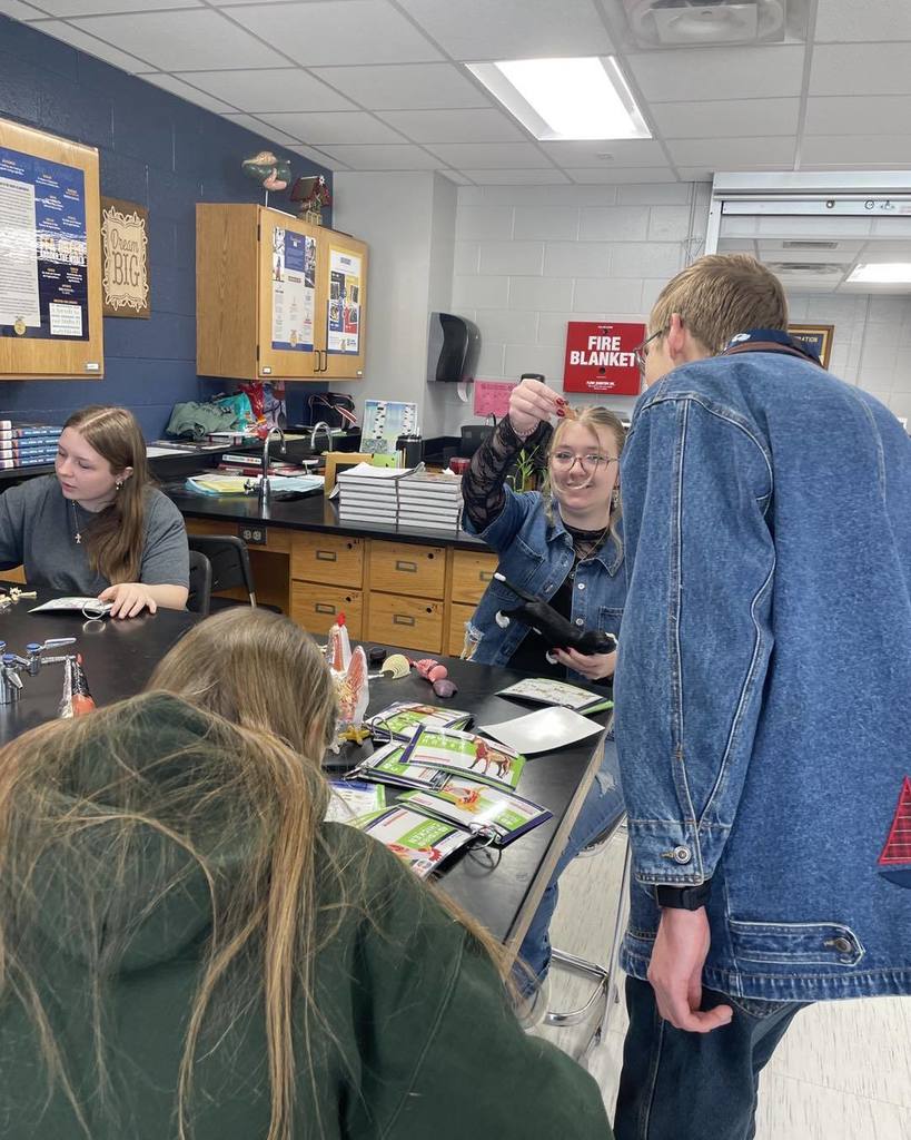 A high school student standing in a science classroom, smiling and holding up a small medical tool or specimen to show her classmates during the vet science lab stations.