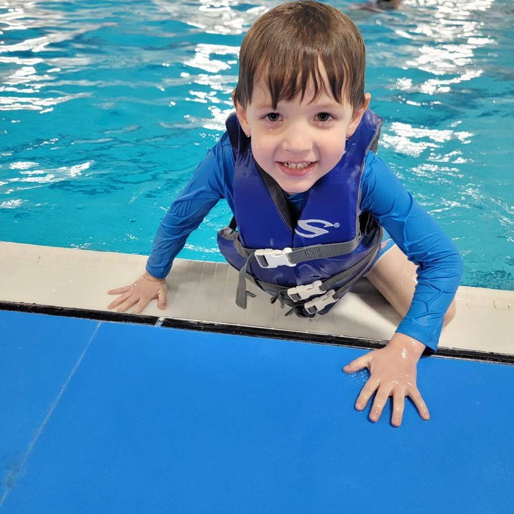 A young student from the Busy Bugs class smiling at the camera while leaning on a blue pool mat at the edge of the school pool. He is wearing a blue long-sleeved swim shirt and a navy blue life jacket.