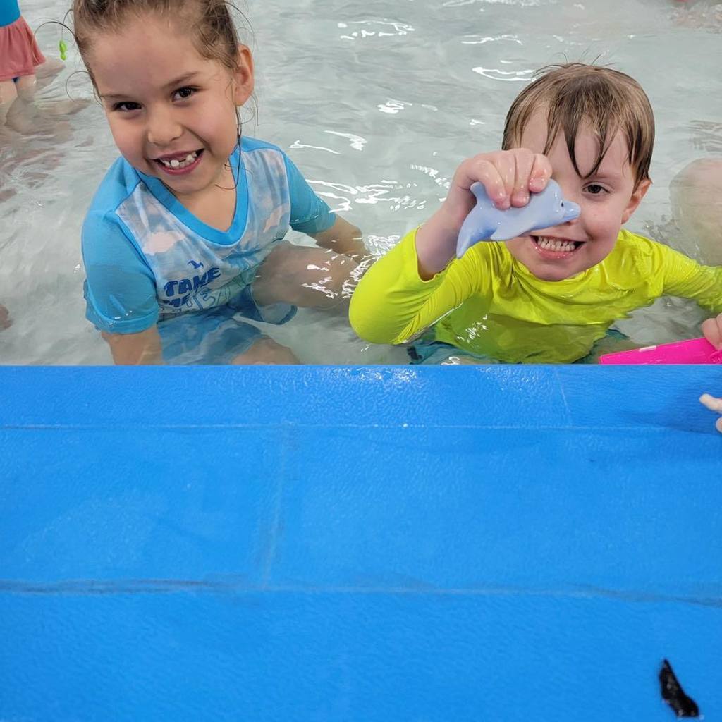 Two young students from the Busy Bugs class smiling in the school pool. One student in a blue and white swim shirt is splashing, while the other in a bright yellow shirt holds up a small blue toy shark.