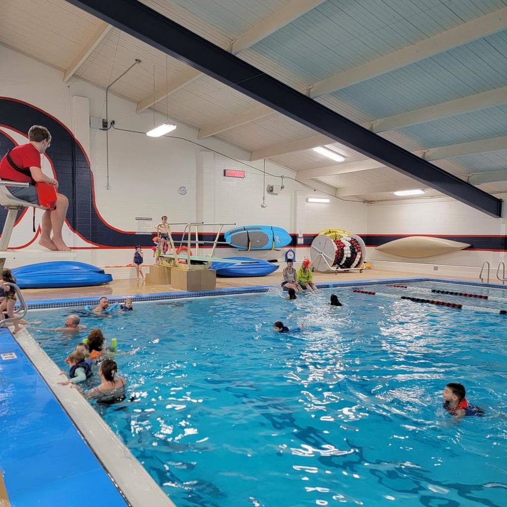 For the final photo in your gallery, which shows the full view of the pool area, here is the alt text: "A wide shot of the Unity school pool during the Busy Bugs ocean unit celebration. Several students and adults are in the water, a lifeguard sits on an elevated chair, and colorful pool floats and equipment are visible along the far wall.