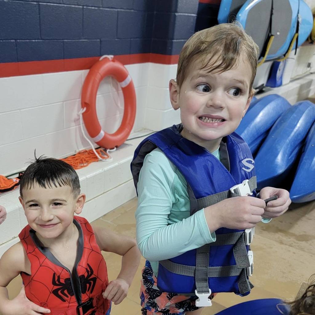 Two young students from the Busy Bugs class posing by the pool during their ocean unit celebration. One student is wearing a red Spider-Man life jacket and the other is wearing a blue life jacket over a teal rash guard.