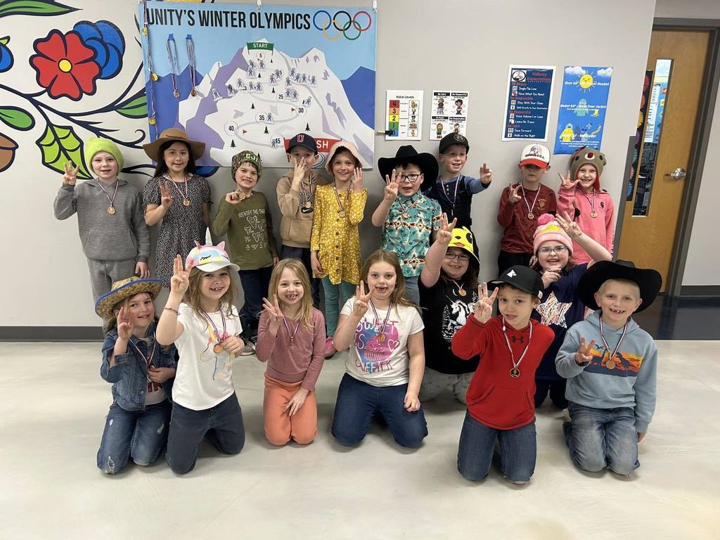A group of Mrs. McElhone’s first-grade students posing together in front of a 'Unity’s Winter Olympics' wall display. The students are smiling and many are wearing cowboy hats and holding up three fingers to represent their 3rd place bronze win.