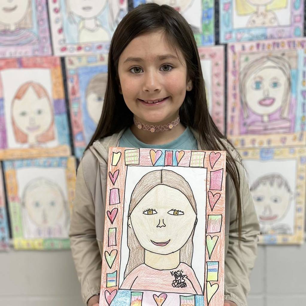 First-grade student Charlotte smiling and holding up her self-portrait drawing. The artwork features a colorful border decorated with hearts and patterns, and is displayed in front of a wall filled with other student portraits.
