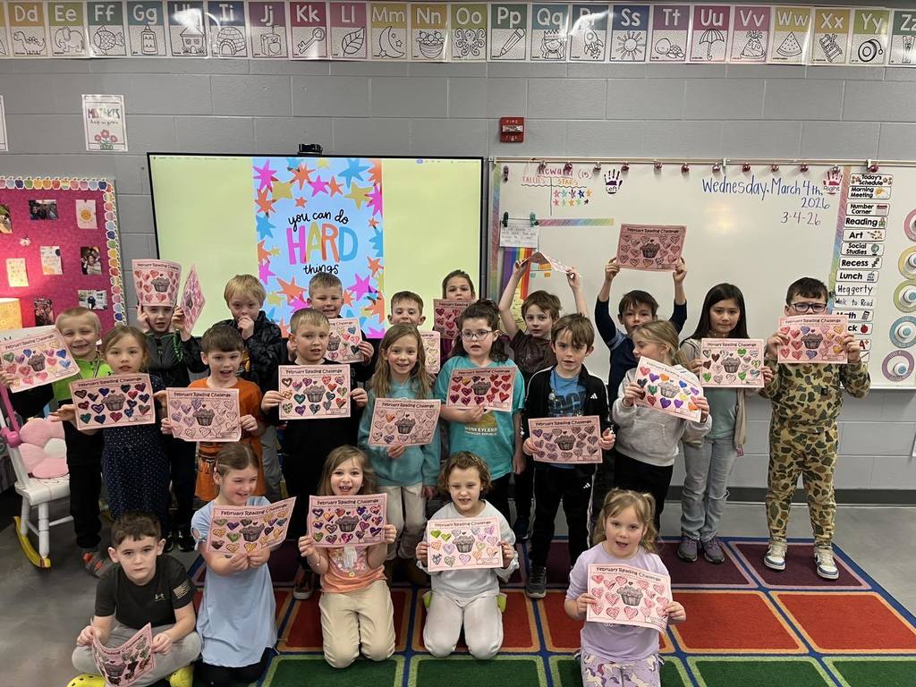 For the group of "February Rockstar Readers," here is a descriptive alt text for your image: "A large group of first-grade students at Unity School District standing together in a classroom. They are proudly holding up their colorful February Reading Challenge certificates, which feature a cupcake design. In the background, a digital screen displays the message 'You can do hard things.