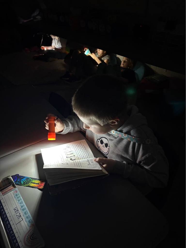 A young student in a grey shirt sitting at a desk in a dark classroom, using a small orange flashlight to read an open book. Other students are visible in the background, also using flashlights to read.