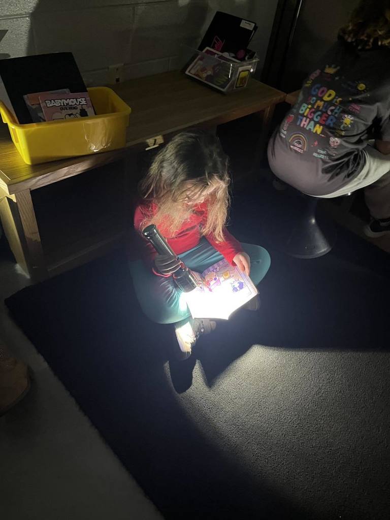A young student with long hair sitting on the floor of a darkened classroom during Flashlight Reading. She is focused on a book in her lap, which is brightly lit by a flashlight she is holding. A yellow bin and other books are visible on a shelf behind her.