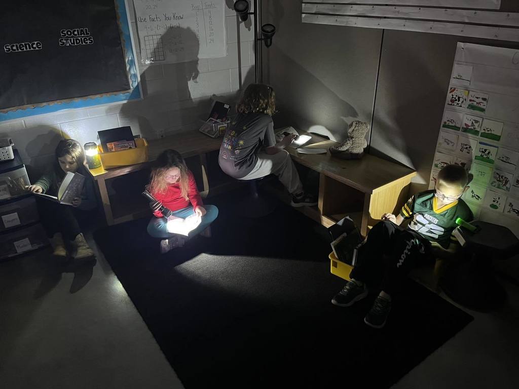 Four third-grade students sitting in a cozy, darkened classroom during Flashlight Reading. They are spread out on the floor and on low benches, each using their own flashlight to light up their books. A 'Science' and 'Social Studies' board is visible on the wall behind them.
