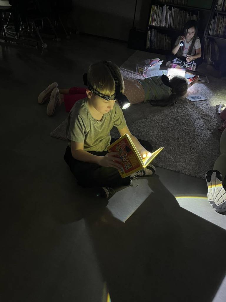 A third-grade student sitting on a classroom rug, wearing a headlamp to read a book during Flashlight Reading. In the background, two other students are lying on the floor, also focused on their books in the dimly lit room.
