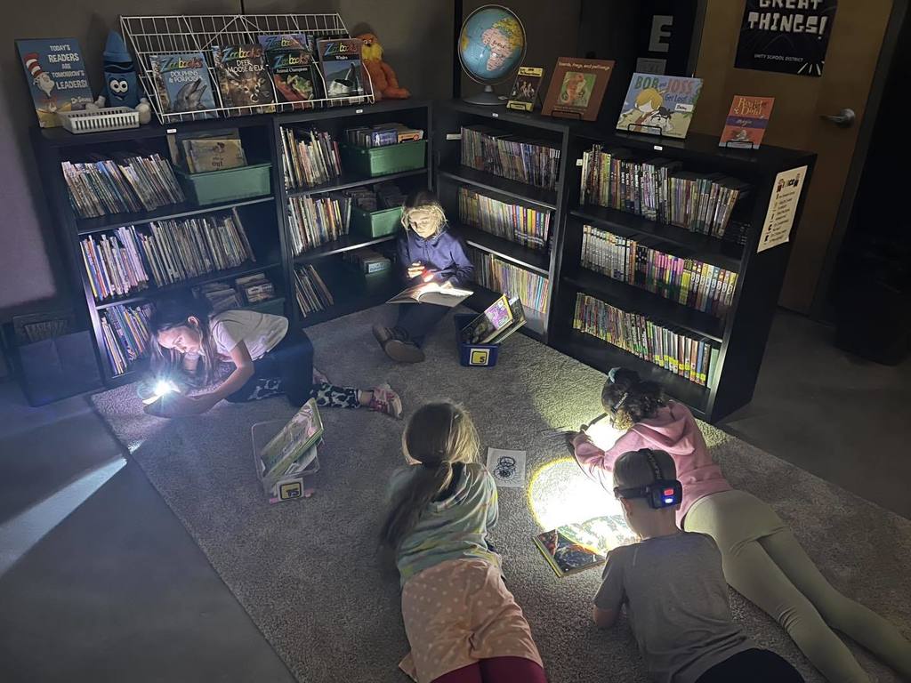 A group of third-grade students gathered in their classroom library during a Flashlight Reading event. The room is dark, and the students are sitting on the floor using flashlights and headlamps to read books. Large bookshelves filled with many books line the walls in the background.