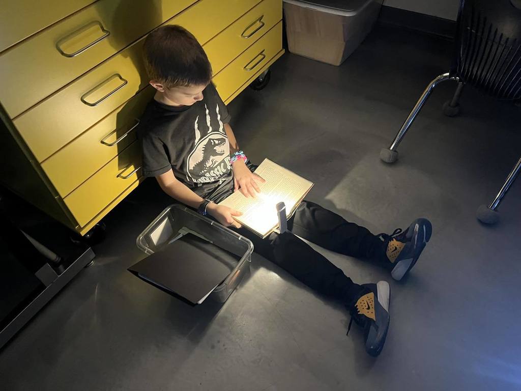 A young student sitting on the floor in a dark classroom, using a small red flashlight to read an open book during Flashlight Reading. The light focuses directly on the pages of the book.