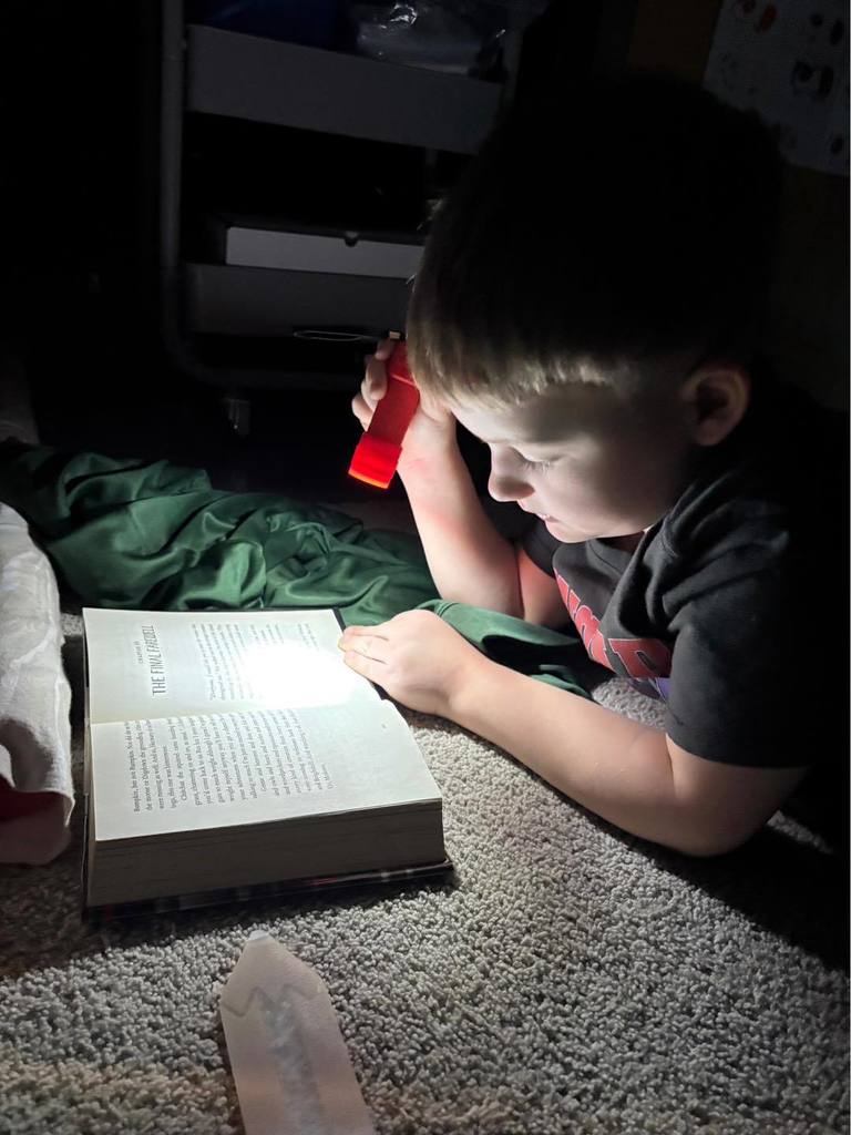 A third-grade student lying on a rug in a dimly lit classroom, using a green flashlight to read. The student is resting their chin on their hands while focused on the pages of an open book.