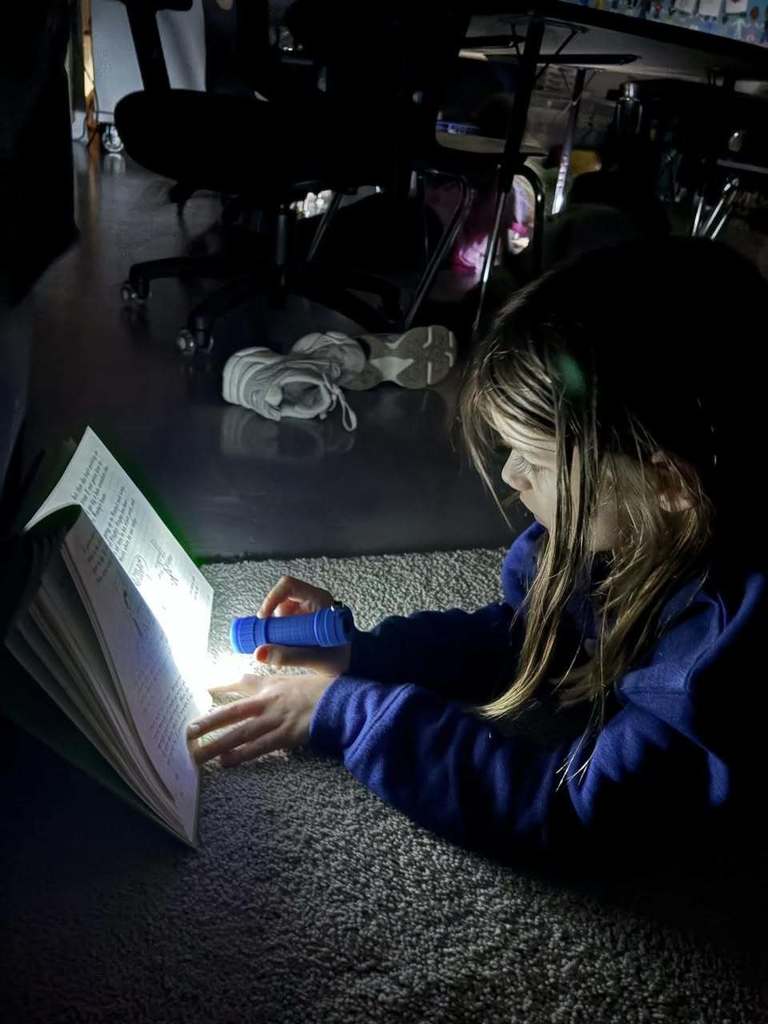 For the fifth photo in your series (the student in the blue sweatshirt), here is the alt text: "A third-grade student in a blue sweatshirt sitting on the floor of a dark classroom, using a small blue flashlight to illuminate the pages of their book during a Read Across America Week activity.