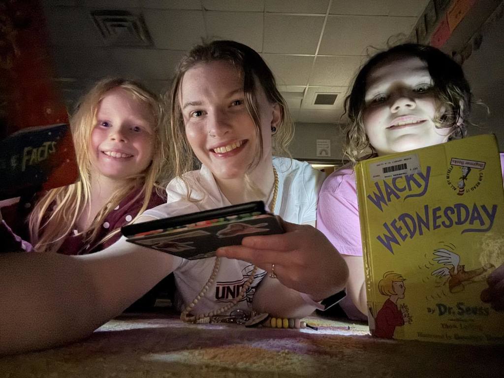 Three smiling third-grade students huddled together on the floor during Flashlight Reading. One student holds up a copy of 'Wacky Wednesday' by Dr. Seuss, which is illuminated by a flashlight in the darkened classroom.