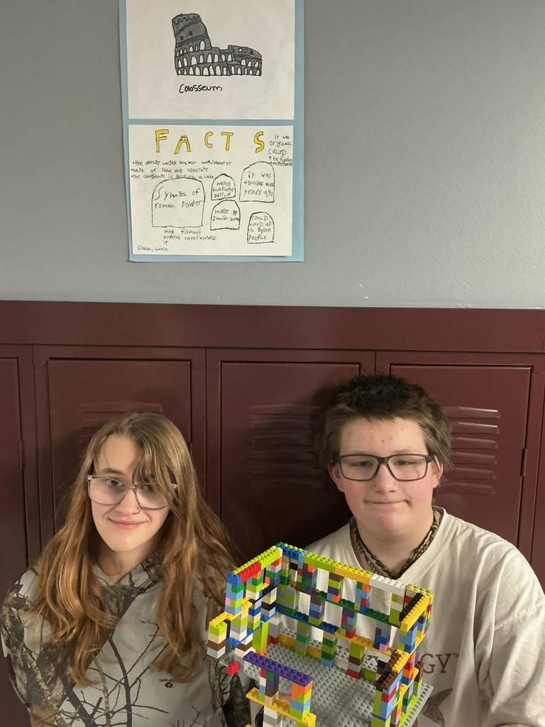 Two middle school students sitting in front of a locker, smiling and posing with their LEGO model of the Roman Colosseum. Above them on the locker is a poster titled 'The Colosseum' featuring facts and a drawing of the landmark.