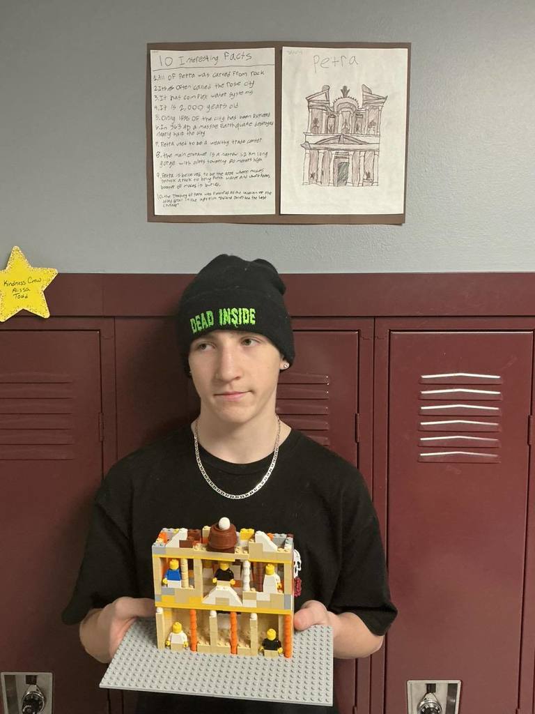 A middle school student wearing a black beanie and sweatshirt, holding a detailed LEGO model of the Treasury at Petra. On the wall behind him are two posters: one titled 'Petra' with a drawing of the landmark, and another listing '10 Interesting Facts' about the site.